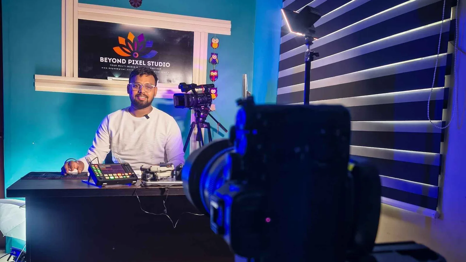 The studio rigged for a podcast — presenter at the desk behind a vision switcher, key light and camera in the foreground, branded backdrop behind