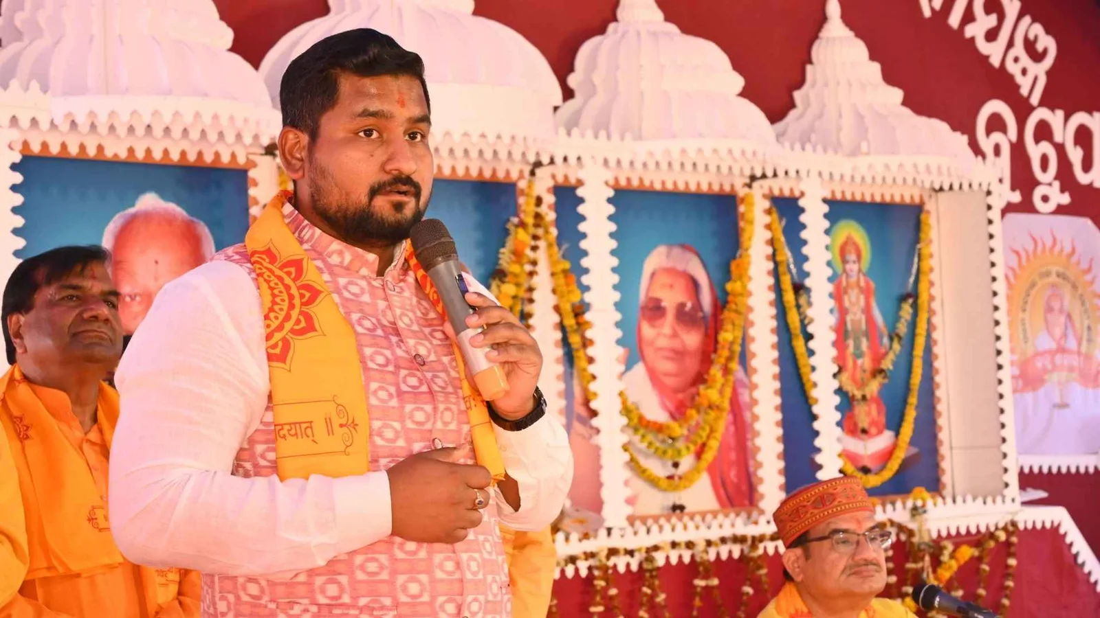 A speaker addressing a garlanded ceremonial event, framed portraits arranged on the stage behind him
