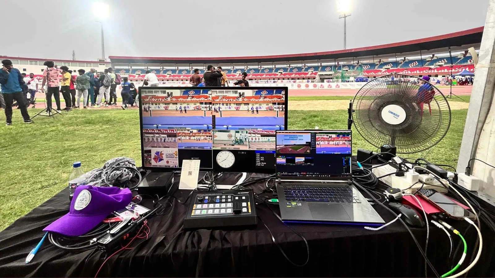Outdoor broadcast desk at trackside — monitors, vision switcher and laptop rigged on a table beside the running track
