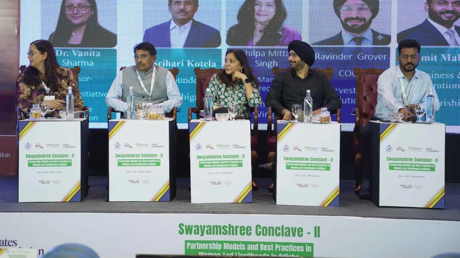 Panel session in progress at Swayamshree Conclave II, speakers seated at the table with their portraits on the screen behind